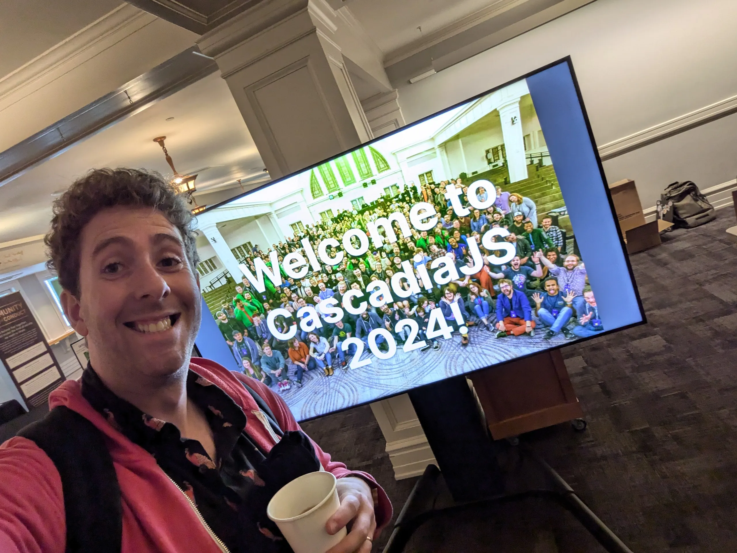 Smiling selfie in front of a TV displaying 'Welcome to CascadiaJS 2024!' in front of a photo.